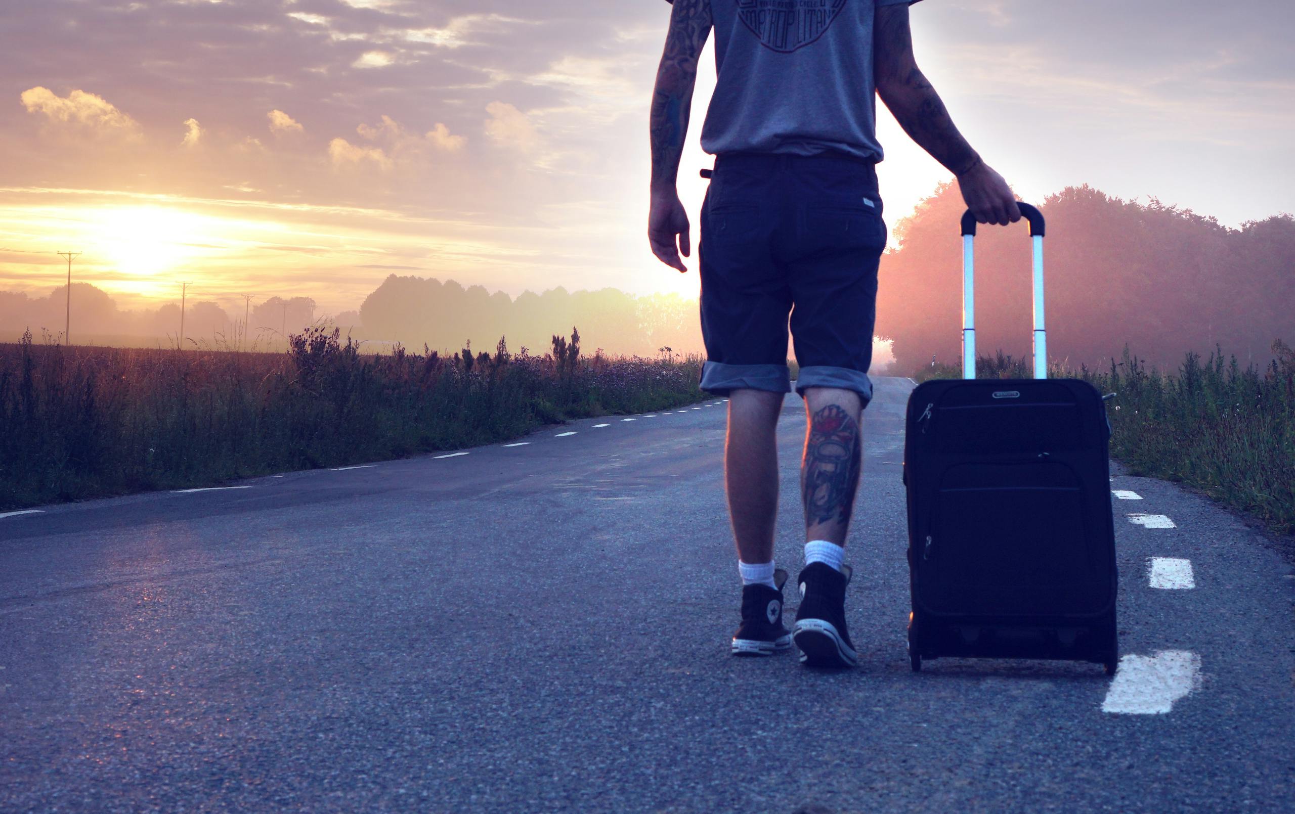 Man with tattoo walks along a rural road at sunrise with a suitcase, showcasing travel and adventure.